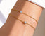 A wrist adorned with two slender gold bracelets the 'Petit Jolie', each with a single sparkling diamond, against a white sleeve.