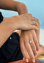 A close-up of a woman's hand adorned with the elegant Jolie ring, featuring a radiant round-cut diamond set in a delicate yellow gold band, against a serene beach backdrop.
