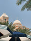 View of a light peach-colored building with dome-shaped roofs, partially obscured by palm trees and blue and white umbrellas. The scene is set under a clear, blue sky.